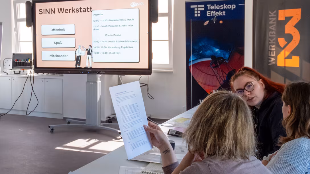 : Drei Personen im Gespräch. Im Hintergrund ist ein großer Screen zu sehen, auf dem „SINN Werkstatt“ zu lesen ist.