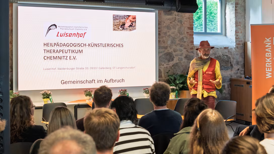 Eine männliche Person mit Hut spricht zum Publikum. Auf dem Screen links neben ihr ist unter anderem zu lesen „Luisenhof. Gemeinschaft im Aufbruch“.