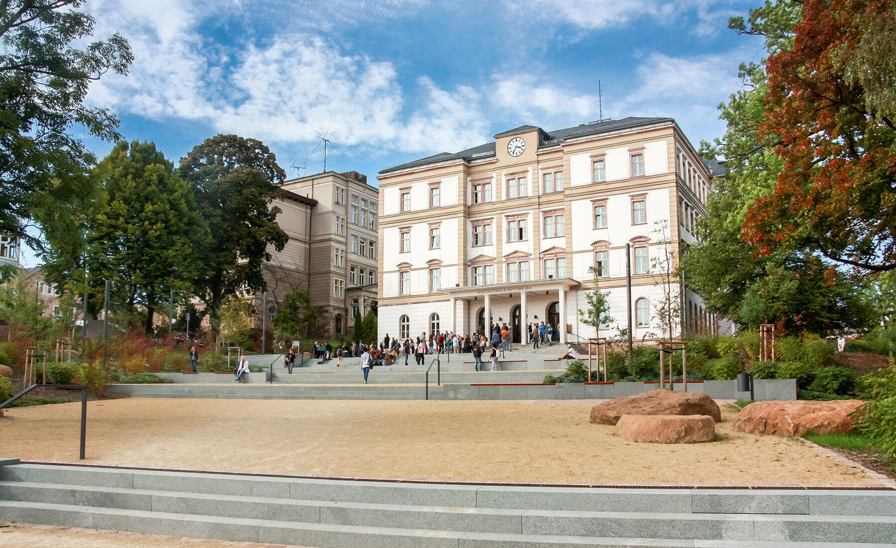 Großes historisches Gebäude mit mehreren Stockwerken, davor ein offener Platz mit Bäumen und Bänken