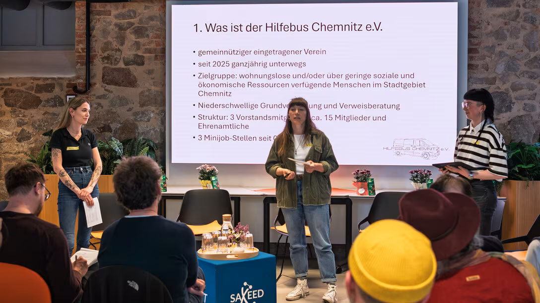 Drei weibliche Personen stehen vor einem Screen. Die mittlere Person spricht zum Publikum. Auf dem Screen ist eine Folie mit der Überschrift „Was ist der Hilfebus Chemnitz e.V.“ und weiteren Anstrichen zu sehen.