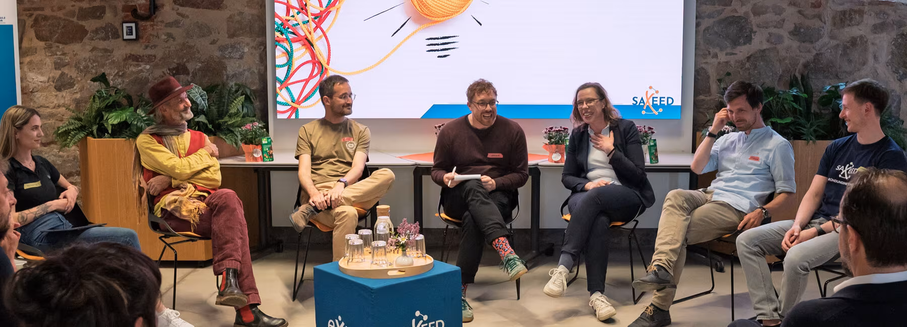 Sieben Personen auf Stühlen vor einem großen Screen, auf dem „Social Entrepreneurship. Wir wird aus sozialem Impact ein tragfähiges Geschäftsmodell“ zu lesen ist. Im Bildvordergrund sind Köpfe von Zuschauenden von hinten zu sehen.
